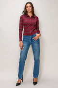 A woman modeling a long-sleeve red and black plaid button-up shirt with blue jeans and black heels, standing against a light gray studio background.