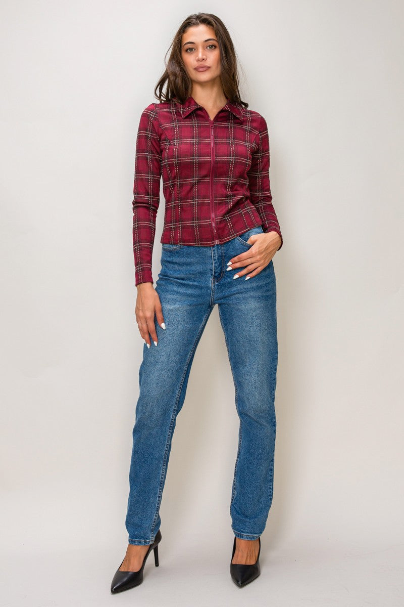 A woman modeling a long-sleeve red and black plaid button-up shirt with blue jeans and black heels, standing against a light gray studio background.