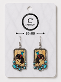 A pair of rectangular dangling earrings displayed on a white retail card. The earrings have silver-tone metal borders and hooks, turquoise blue beads, and a central printed graphic depicting a cowboy riding a bucking horse.