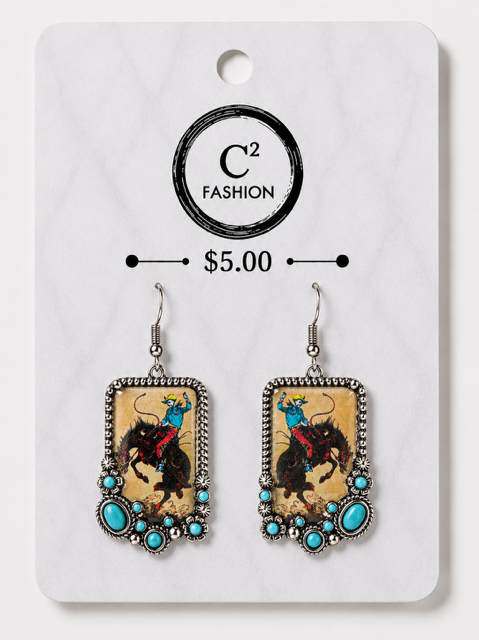 A pair of rectangular dangling earrings displayed on a white retail card. The earrings have silver-tone metal borders and hooks, turquoise blue beads, and a central printed graphic depicting a cowboy riding a bucking horse.