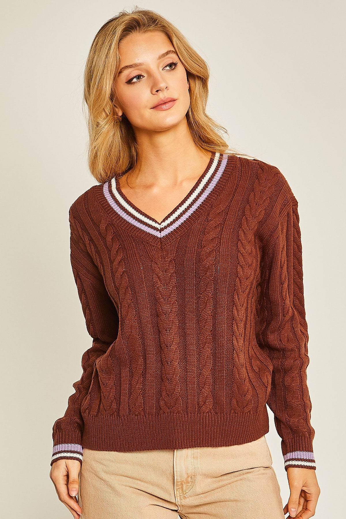 A woman wearing a brown cable-knit V-neck sweater with striped trim in white and purple, paired with beige pants, against a light beige studio background.