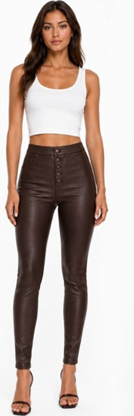 A woman modeling high-waisted, dark brown faux-leather pants with a button fly, paired with a white crop top and black heels, against a plain white background.