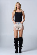 A woman stands against a neutral gray background wearing a fitted black spaghetti-strap top paired with high-waisted distressed denim shorts. The shorts feature a light-to-beige wash with heavy distressing on the front and frayed hems. She completes the look with tall, black slouchy boots, giving the outfit a bold, trendy vibe.