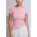 A woman wearing a pink short-sleeve top with white polka dots and a draped neckline, paired with a white skirt, shown against a plain light gray background.