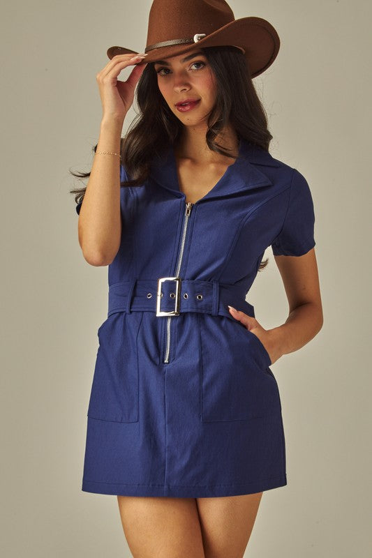 Woman wearing a blue dress with a belt and a brown hat against a neutral background