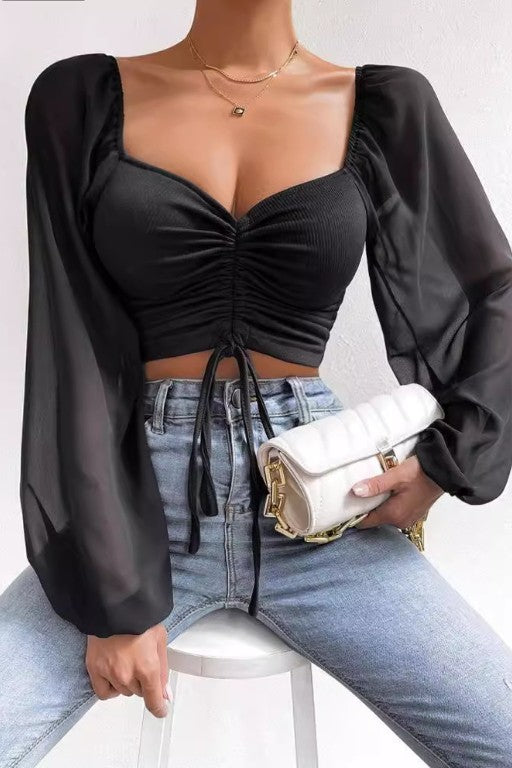 A woman seated on a white stool wearing a black ruched sweetheart crop top with sheer puff sleeves, paired with light blue jeans and holding a white quilted clutch bag.
