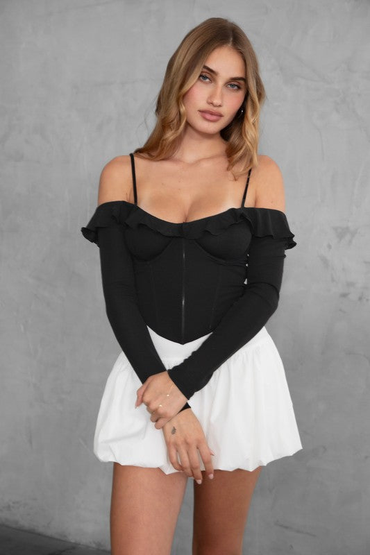 Woman wearing a black off-shoulder top with ruffles and a white skirt against a gray background