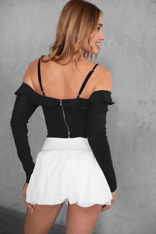 Woman wearing a black off-shoulder top with long sleeves and white shorts against a gray background