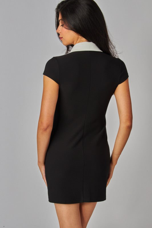 Back view of a woman wearing a fitted black mini dress with short sleeves and a contrasting white collar. The dress features a clean, tailored silhouette with a center back seam and a smooth, minimalist design. She is standing slightly turned to the side against a light gray studio background, showcasing the back fit and structure of the garment.