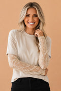A smiling blonde woman wearing a cream-colored short-sleeve top with sheer lace long sleeves, paired with black pants, against a solid peach-colored background.
