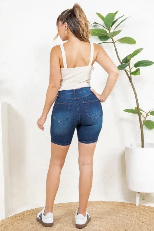 Woman wearing blue denim shorts standing in a room with a plant and white wall.