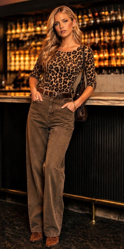 A woman posing in a bar setting wearing a brown leopard-print top, brown wide-leg jeans, and brown boots, with a dark shoulder bag.