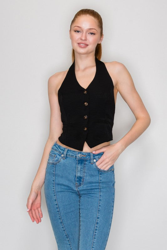 A woman modeling a black halter-neck vest top with front buttons, paired with blue jeans, against a light gray studio background.