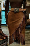 A brown faux-suede fringe skirt with a diagonal wrap-style drape and long fringe hem, styled with a matching belt featuring a large western-style buckle. The skirt is displayed on a mannequin torso in a rustic shop setting.