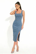 A woman modeling a sleeveless blue denim midi dress with a square neckline, front button detail, and a high side slit, paired with silver high-heeled sandals against a white studio background.
