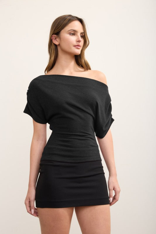A woman modeling a black off-the-shoulder mini dress with short puff sleeves and a ruched bodice, photographed against a plain light beige studio background.