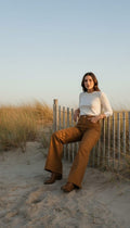 A woman sits on a wooden fence at a dune field during golden hour, wearing a white long-sleeve top and brown wide-leg pants with brown boots.