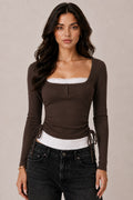model wearing a fitted dark brown ribbed long-sleeve top with a square neckline, button-front detail, and adjustable side ties, layered over a white lace-trim camisole and paired with black patterned jeans.