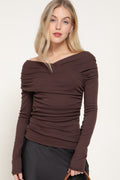 Model wearing a fitted chocolate brown long-sleeve top with an off-the-shoulder neckline and ruched detailing, paired with a black skirt.