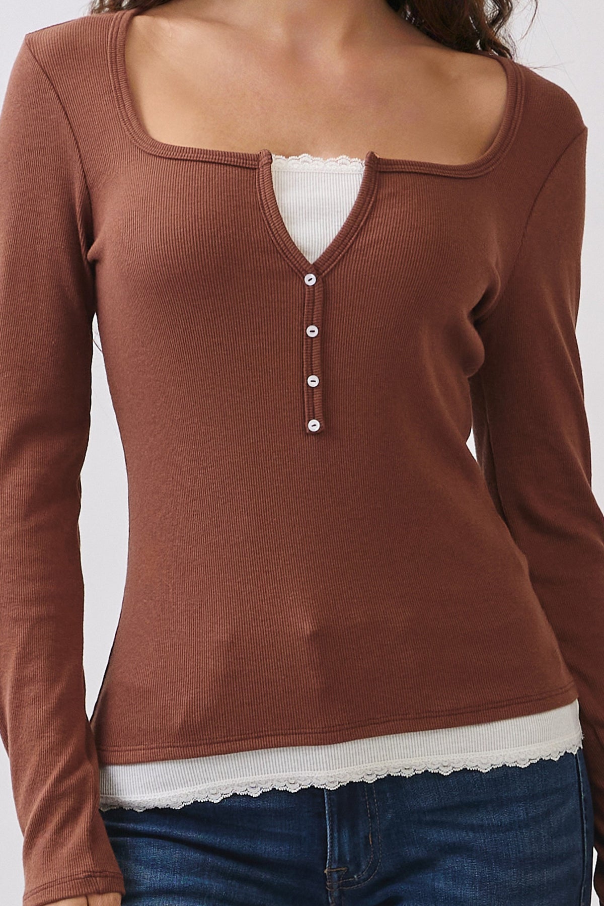 A woman wearing a brown long-sleeve henley top with a scoop neckline, three front buttons, and a white lace-trim hem, paired with blue jeans.