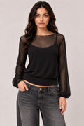 A woman modeling a black sheer long-sleeve top with a scoop neckline over a black camisole, paired with gray jeans, against a solid light gray studio background.