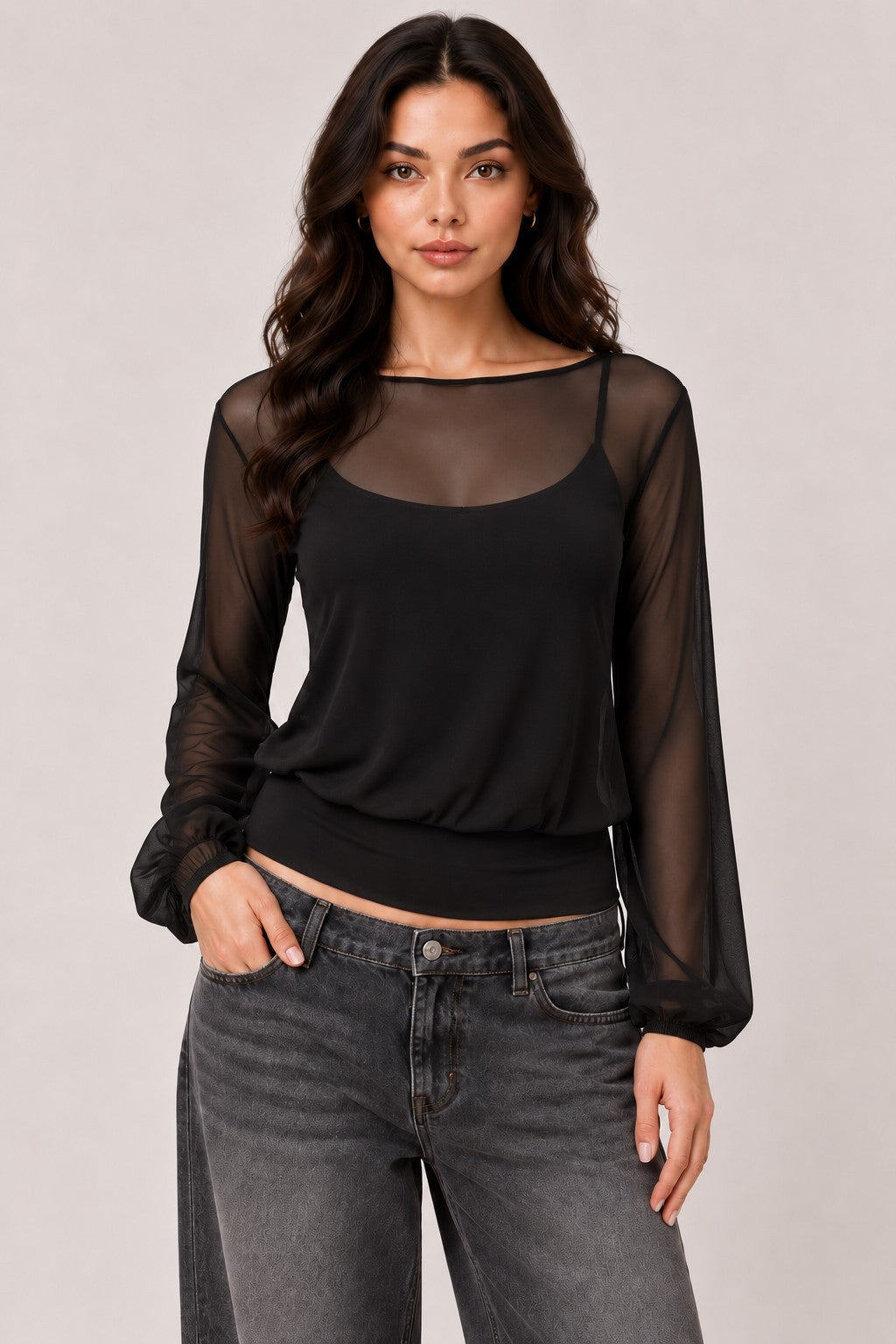 A woman modeling a black sheer long-sleeve top with a scoop neckline over a black camisole, paired with gray jeans, against a solid light gray studio background.