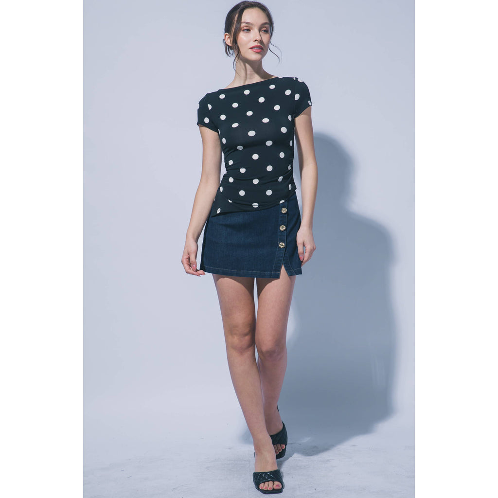 A woman modeling a short-sleeve dark navy dress with white polka dots on the top and a dark denim mini skirt with gold-tone buttons, set against a light gray studio background.