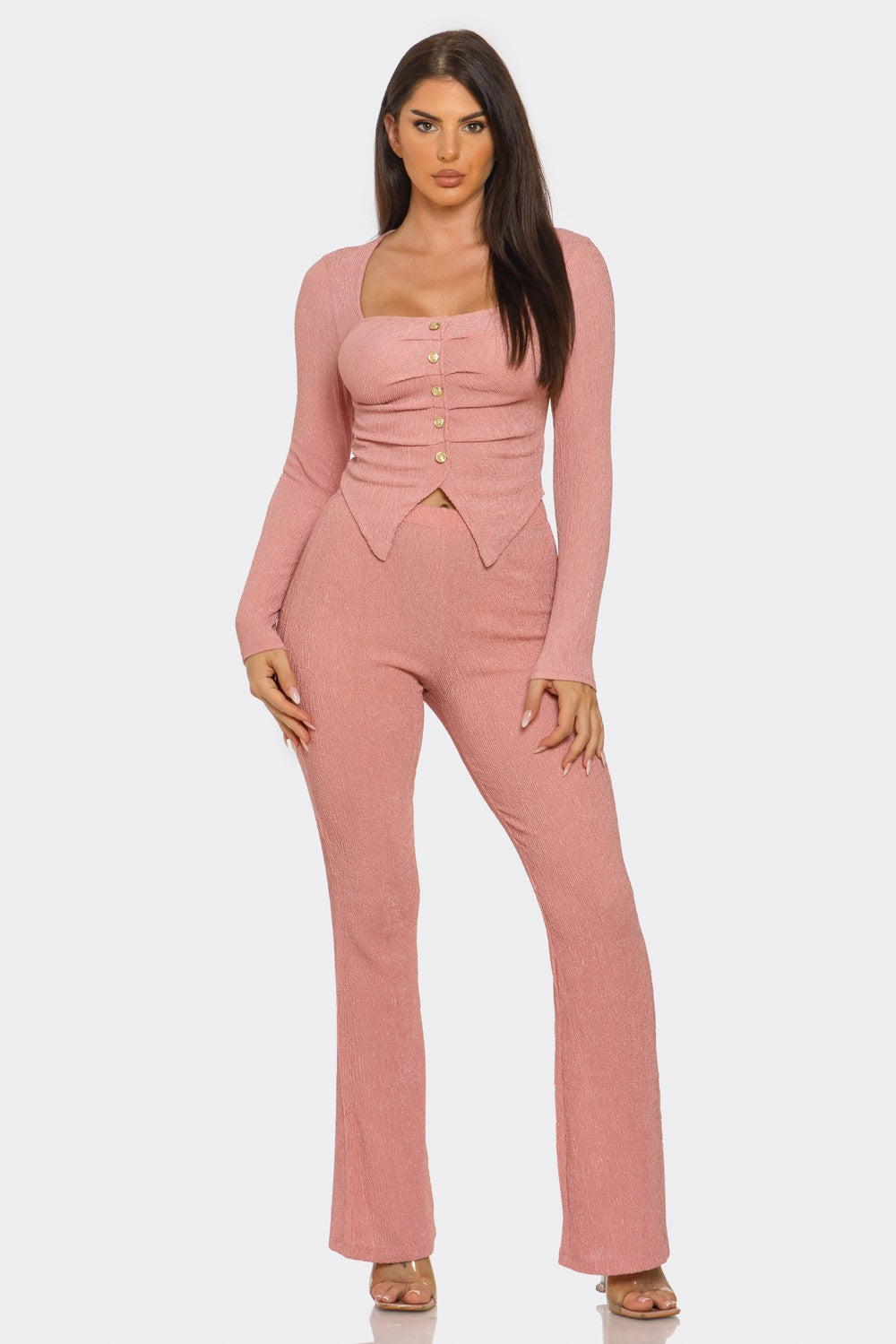 A woman modeling a dusty pink long-sleeve jumpsuit with a structured sweetheart neckline, buttoned corset-style waist, and straight-leg pants, set against a plain light gray studio background.