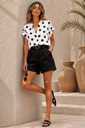 A woman models a white short-sleeve button-up blouse with black polka dots and black high-waisted shorts, styled with a black belt, sunglasses, and black slide sandals, leaning against a textured beige stucco wall.