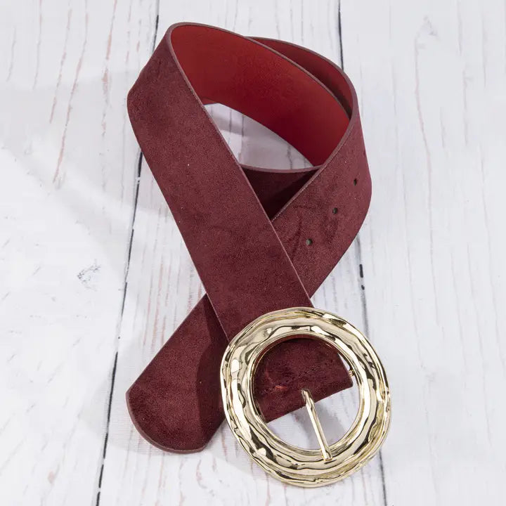 A wide burgundy suede belt with a large, textured gold-tone circular buckle, coiled on a white wooden plank surface.
