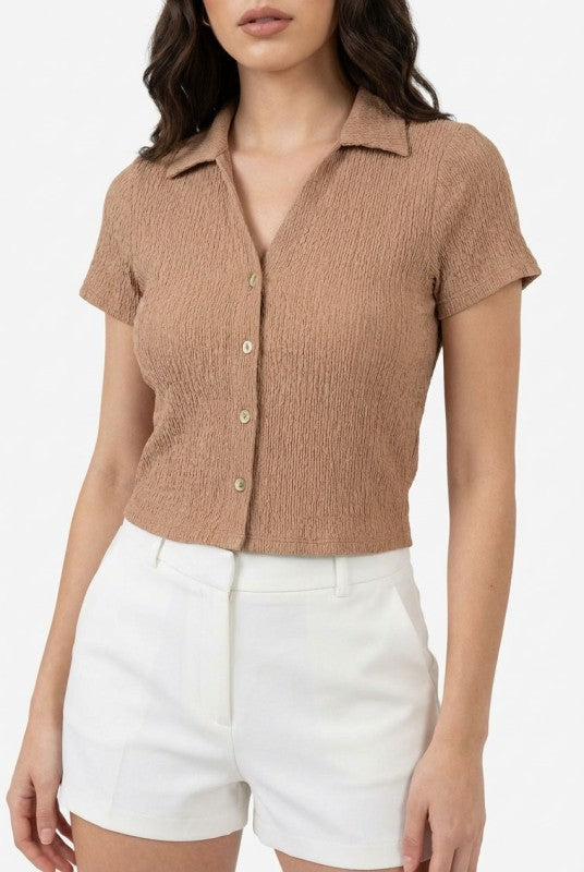 Woman wearing a brown ribbed polo shirt and white shorts on a light gray background.