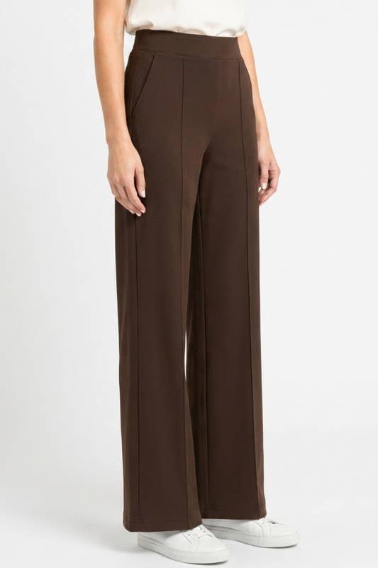 A model wearing high-waisted, wide-leg brown trousers with a white top and white sneakers, shown from the waist down against a light gray studio background.