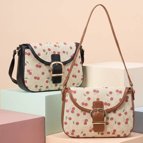Two cherry-patterned shoulder bags displayed on pastel geometric blocks. One bag has black trim and a black buckle closure, while the other has brown trim and a brown buckle closure.