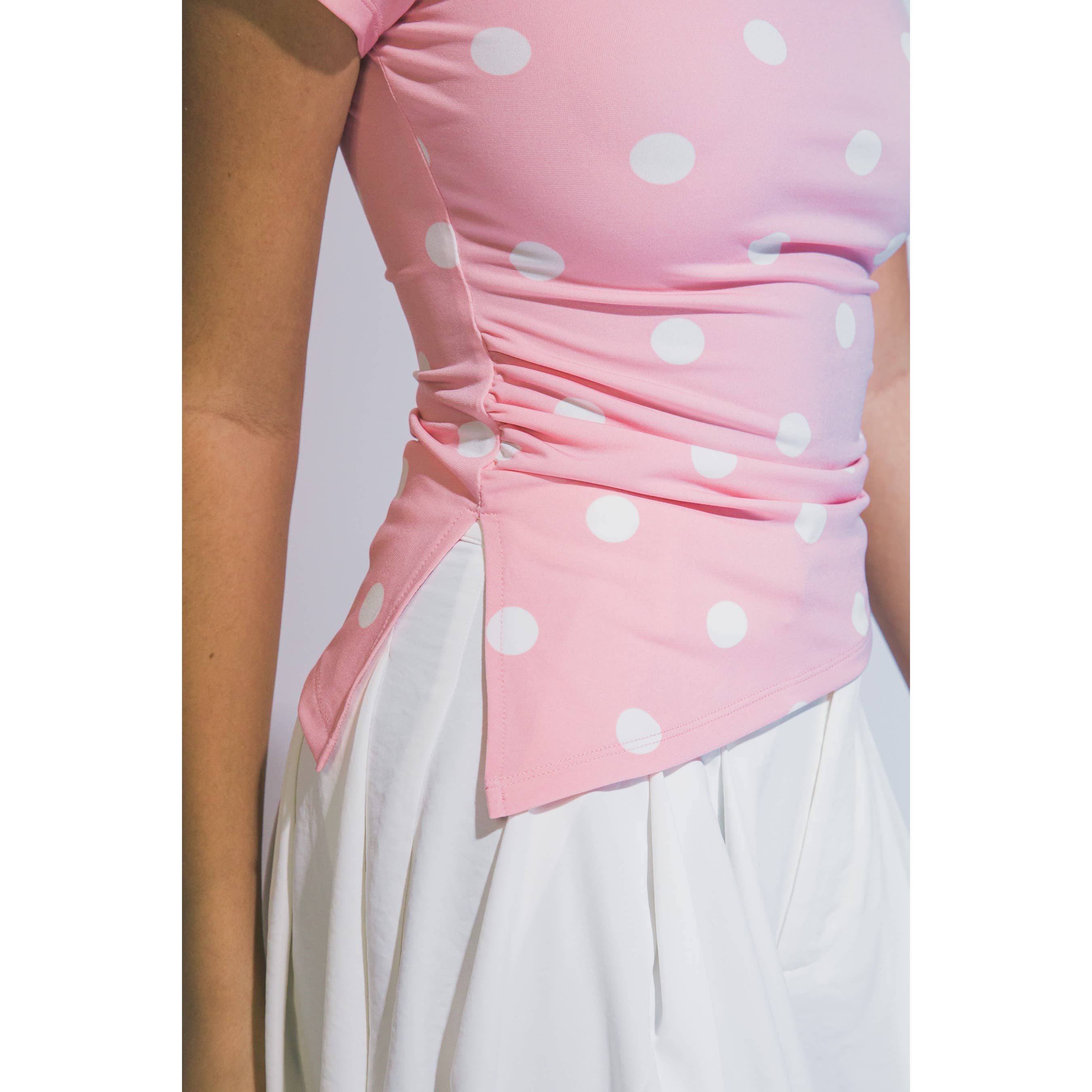 A close-up of a pink sleeveless top with white polka dots and gathered ruching at the waist, worn with a white skirt against a plain light background.