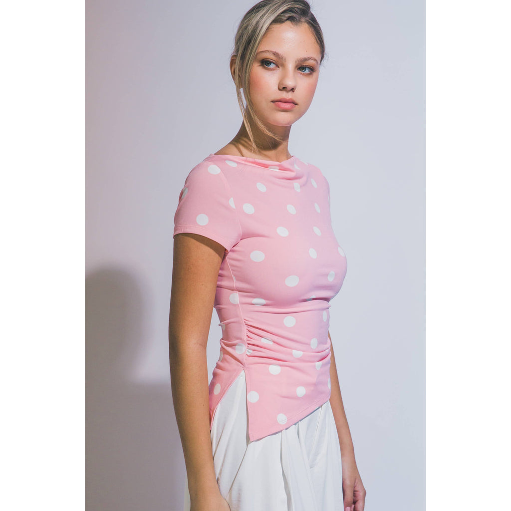 A woman modeling a pink short-sleeve top with white polka dots and a gathered, ruched waist, paired with white bottoms, set against a plain light gray studio background.