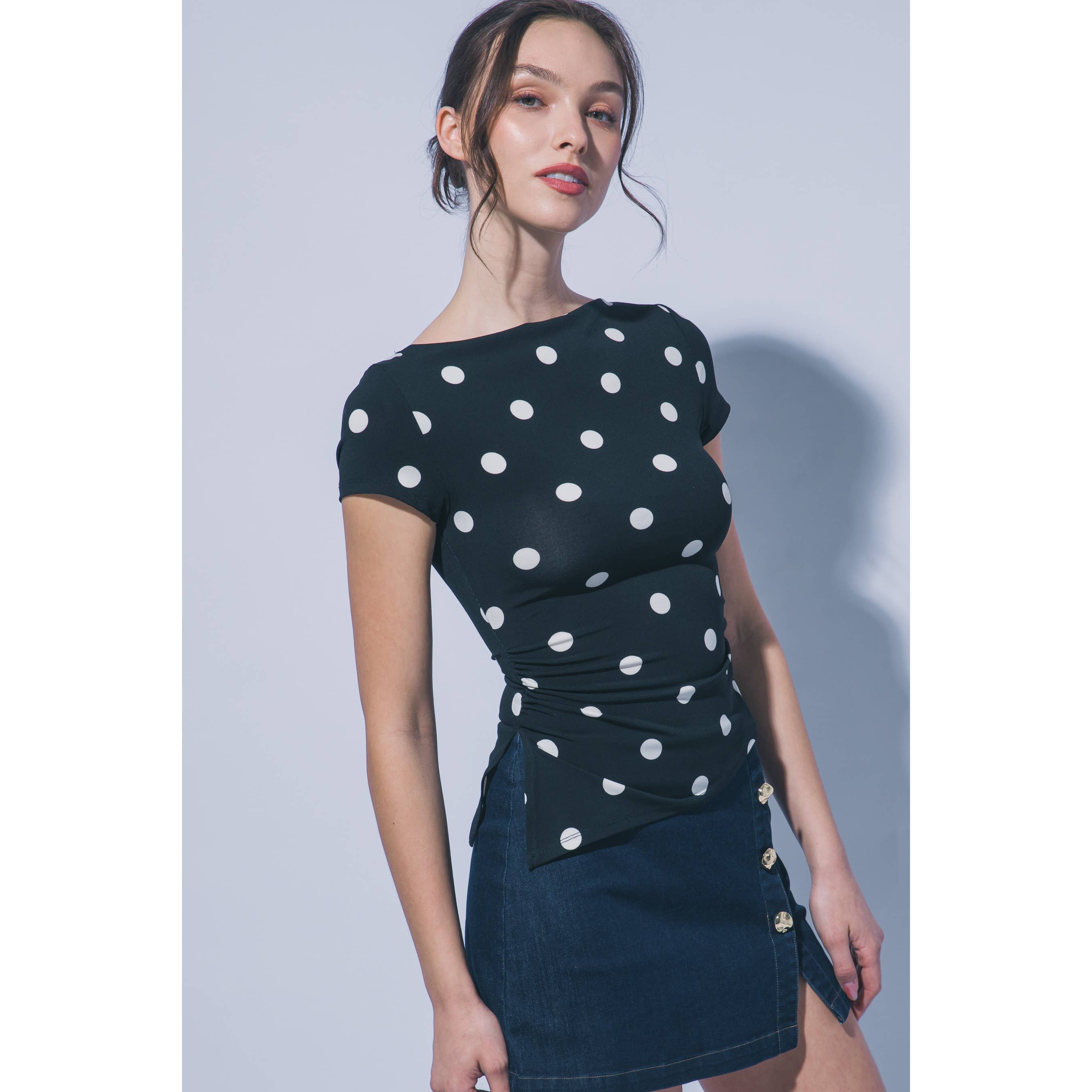 A woman modeling a fitted short-sleeve top and matching mini skirt set. The top is black with large white polka dots, and the skirt is dark blue denim with gold-tone buttons.