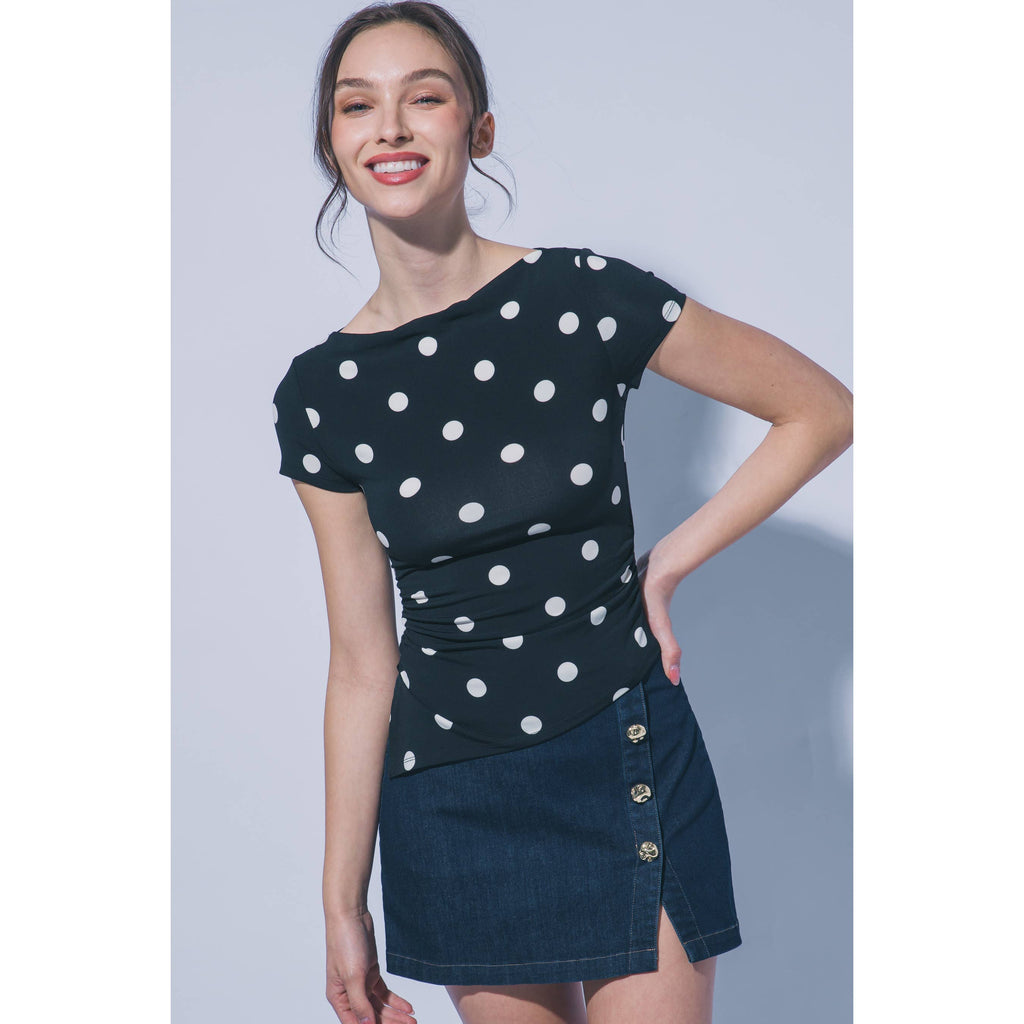 A woman modeling a short-sleeve black top with white polka dots and a dark denim mini skirt with gold-tone buttons, set against a plain light gray studio background.