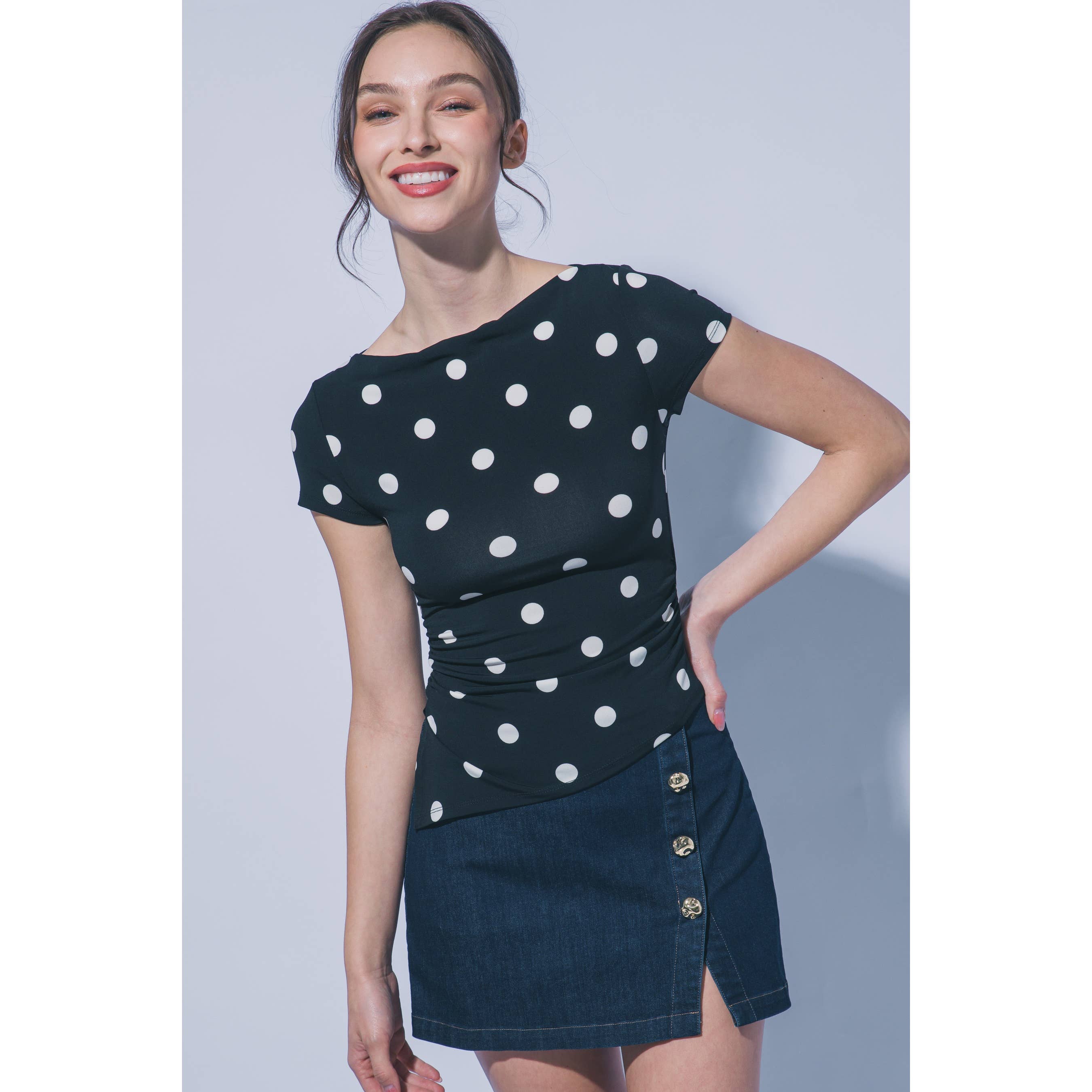 A woman modeling a short-sleeve black top with white polka dots and a dark denim mini skirt with gold-tone buttons, set against a plain light gray studio background.