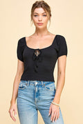 A woman modeling a black short-sleeve top with a square neckline, puff sleeves, and a front tie detail, paired with light blue jeans against a solid beige background.