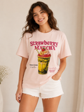 A woman wearing a light pink graphic t-shirt with a 'Strawberry Matcha' drink illustration and white denim shorts, standing against a neutral cream-colored background.
