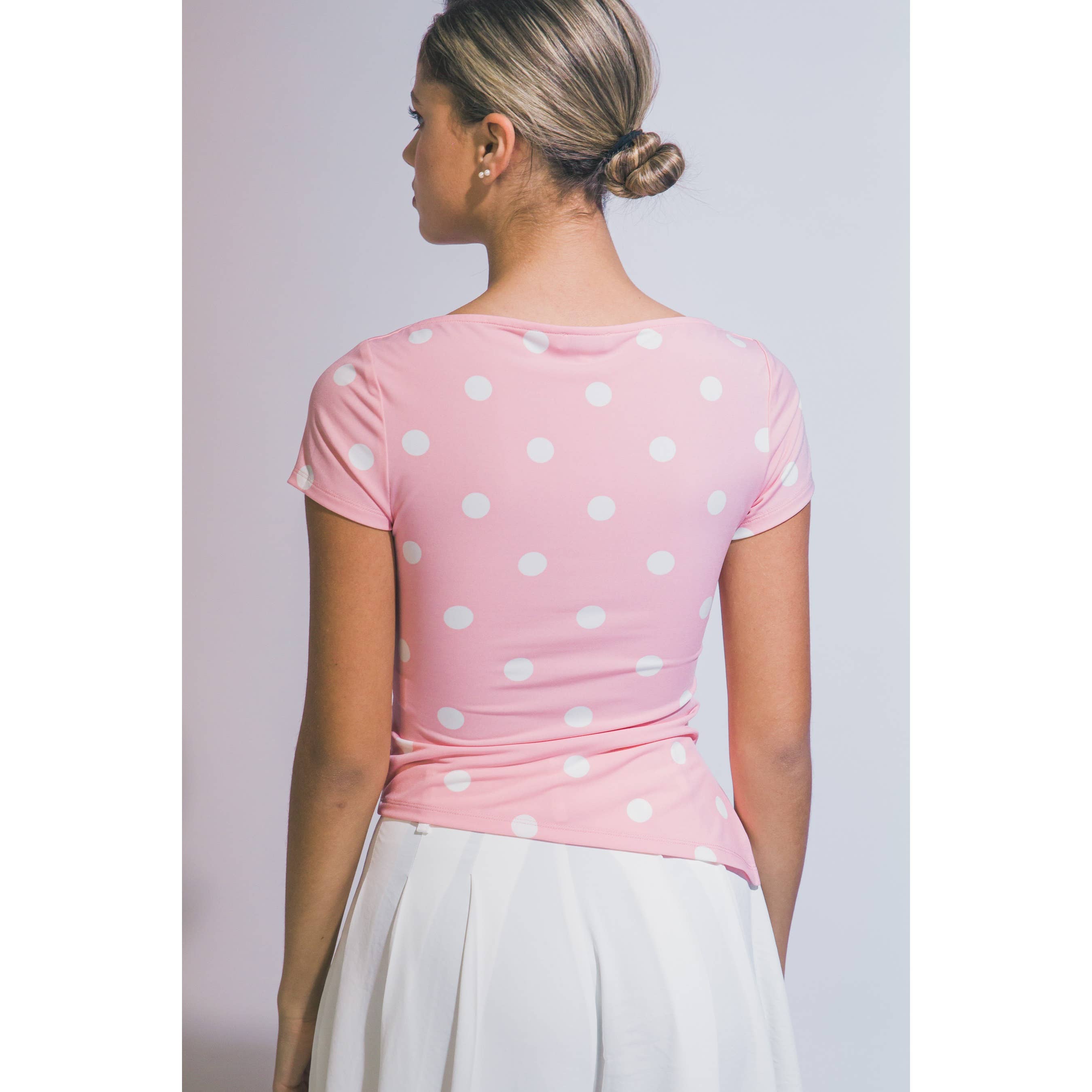 A woman shown from the back wearing a light pink short-sleeve top with white polka dots and a white skirt, set against a plain light gray studio background.