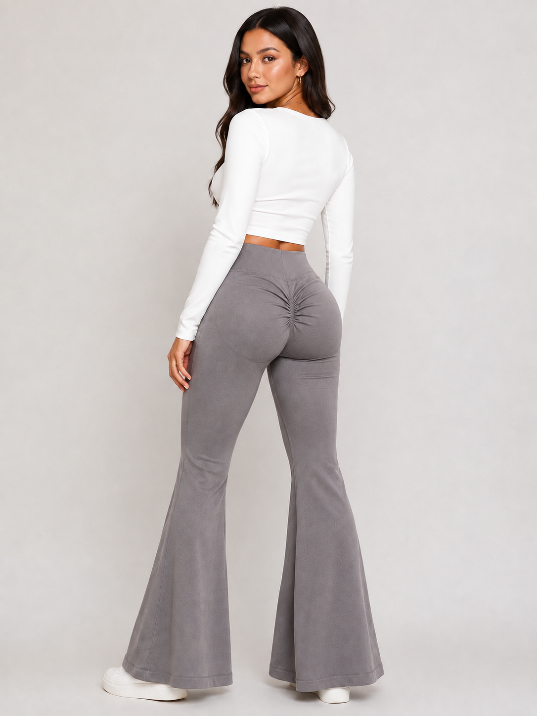A woman modeling high-waisted, gray flared pants with a ruched back detail, paired with a white long-sleeve crop top, shown from a rear three-quarter angle against a light gray studio background.