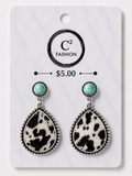 A pair of dangle earrings displayed on a white retail card. Each earring has a round turquoise-colored stone at the top and a large teardrop-shaped black-and-white cow-print drop below, set in silver-tone metal with a beaded border.