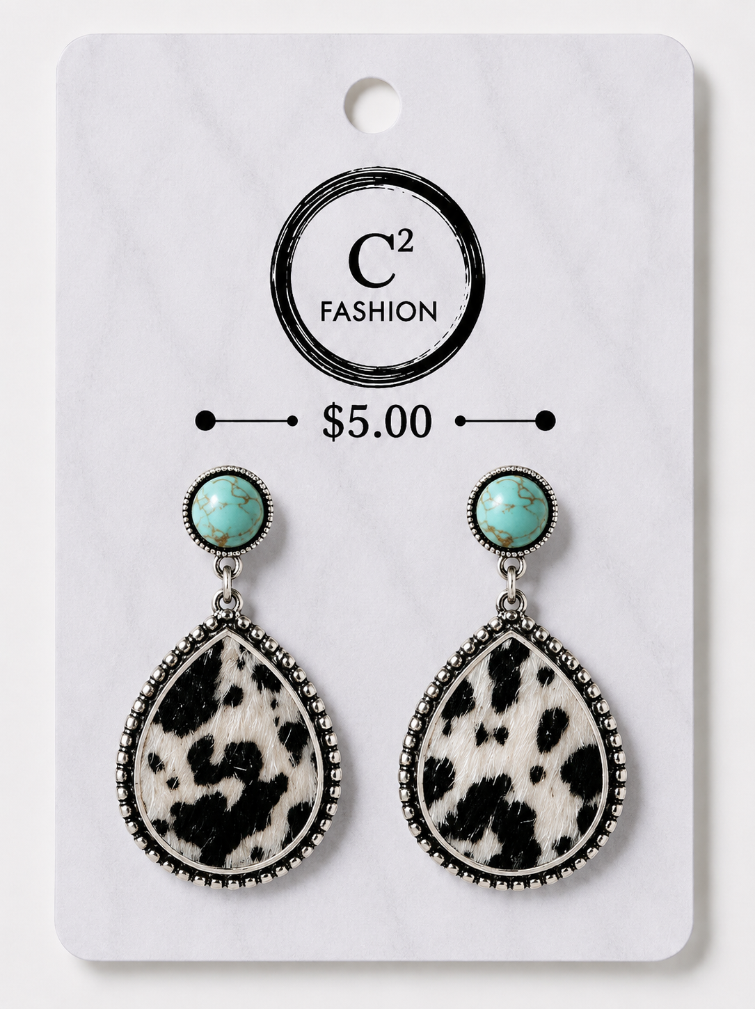 A pair of dangle earrings displayed on a white retail card. Each earring has a round turquoise-colored stone at the top and a large teardrop-shaped black-and-white cow-print drop below, set in silver-tone metal with a beaded border.