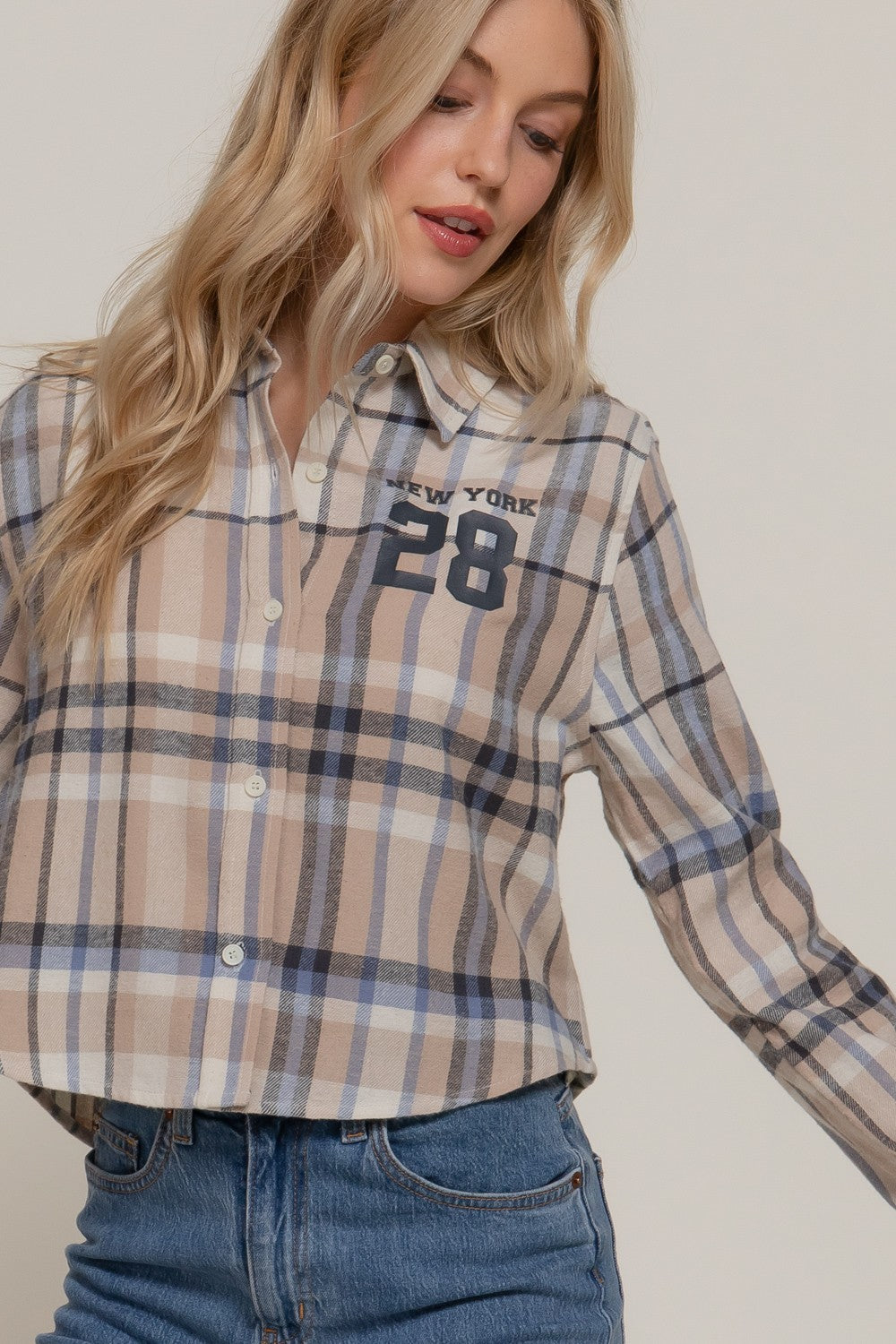 Model wearing a beige plaid button-up shirt with blue and black stripes, white buttons, and a navy “New York 28” graphic on the chest, paired with blue jeans.