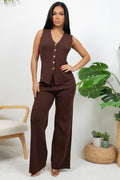 A woman modeling a sleeveless brown vest and matching wide-leg pants set, standing in a bright indoor setting with a chair and potted plants visible.