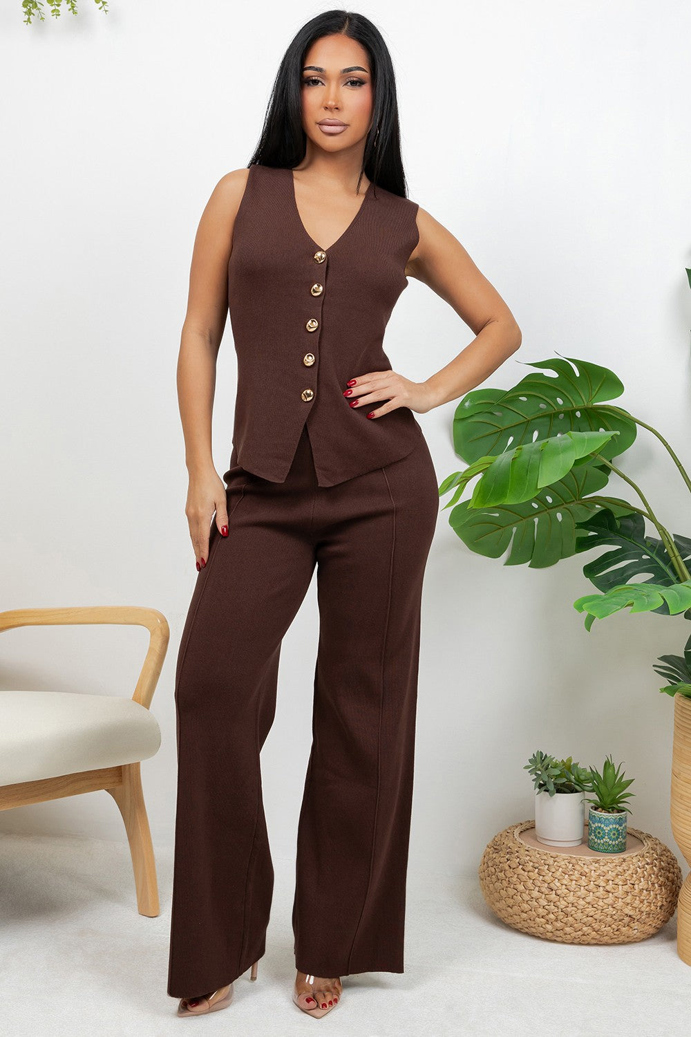 A woman modeling a sleeveless brown vest and matching wide-leg pants set, standing in a bright indoor setting with a chair and potted plants visible.