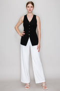 A woman modeling a sleeveless black knit vest with a V-neck and front buttons, paired with white wide-leg pants and white strappy heels, against a light gray studio background.