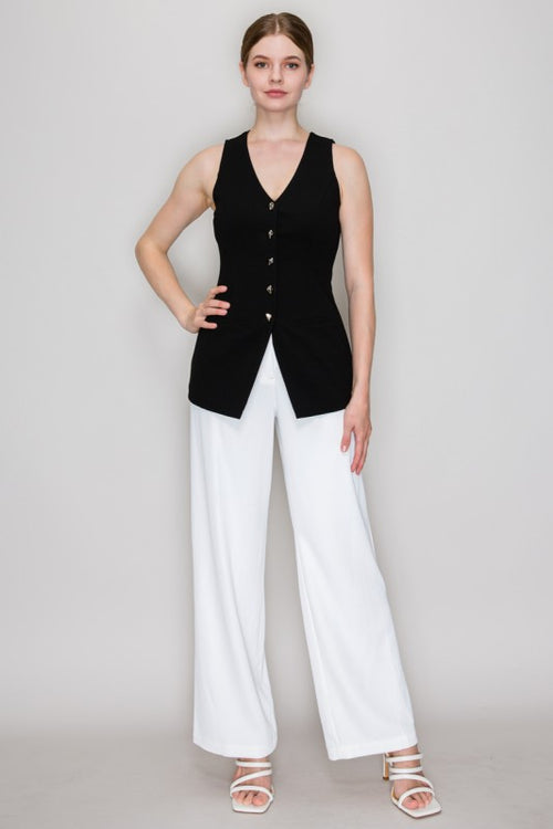 A woman modeling a sleeveless black knit vest with a V-neck and front buttons, paired with white wide-leg pants and white strappy heels, against a light gray studio background.