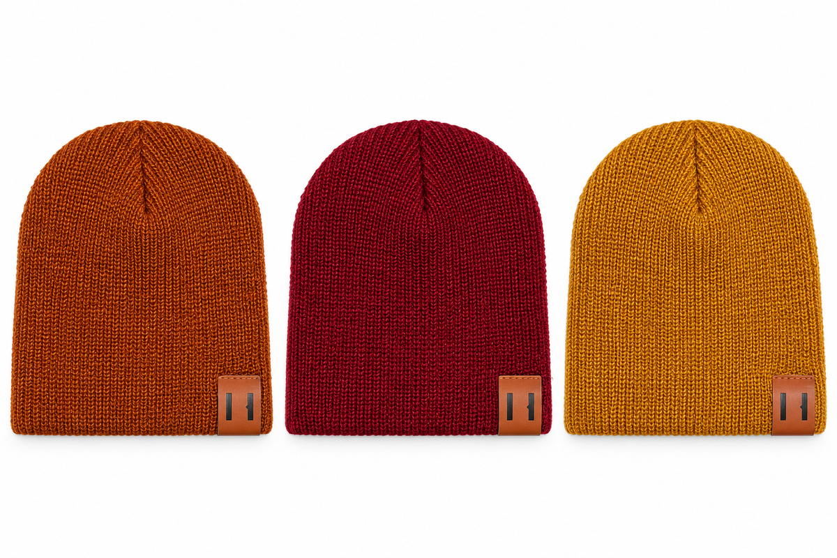 Three ribbed knit beanie hats shown side by side on a plain white background, in rust orange, deep red, and mustard yellow, each with a small tan leather patch near the lower edge.